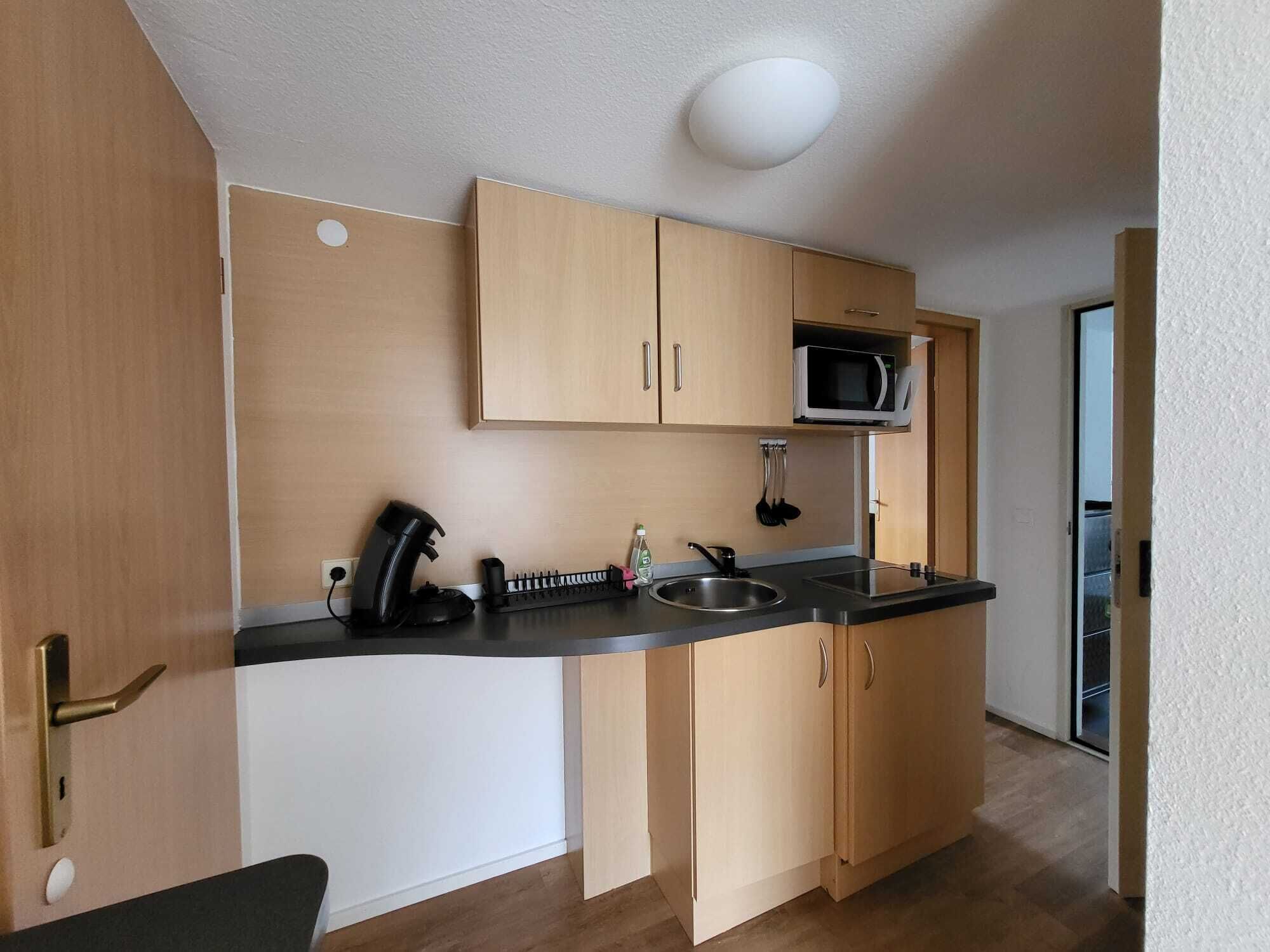 Cocina compacta con armarios de madera, microondas, hervidor y pileta.