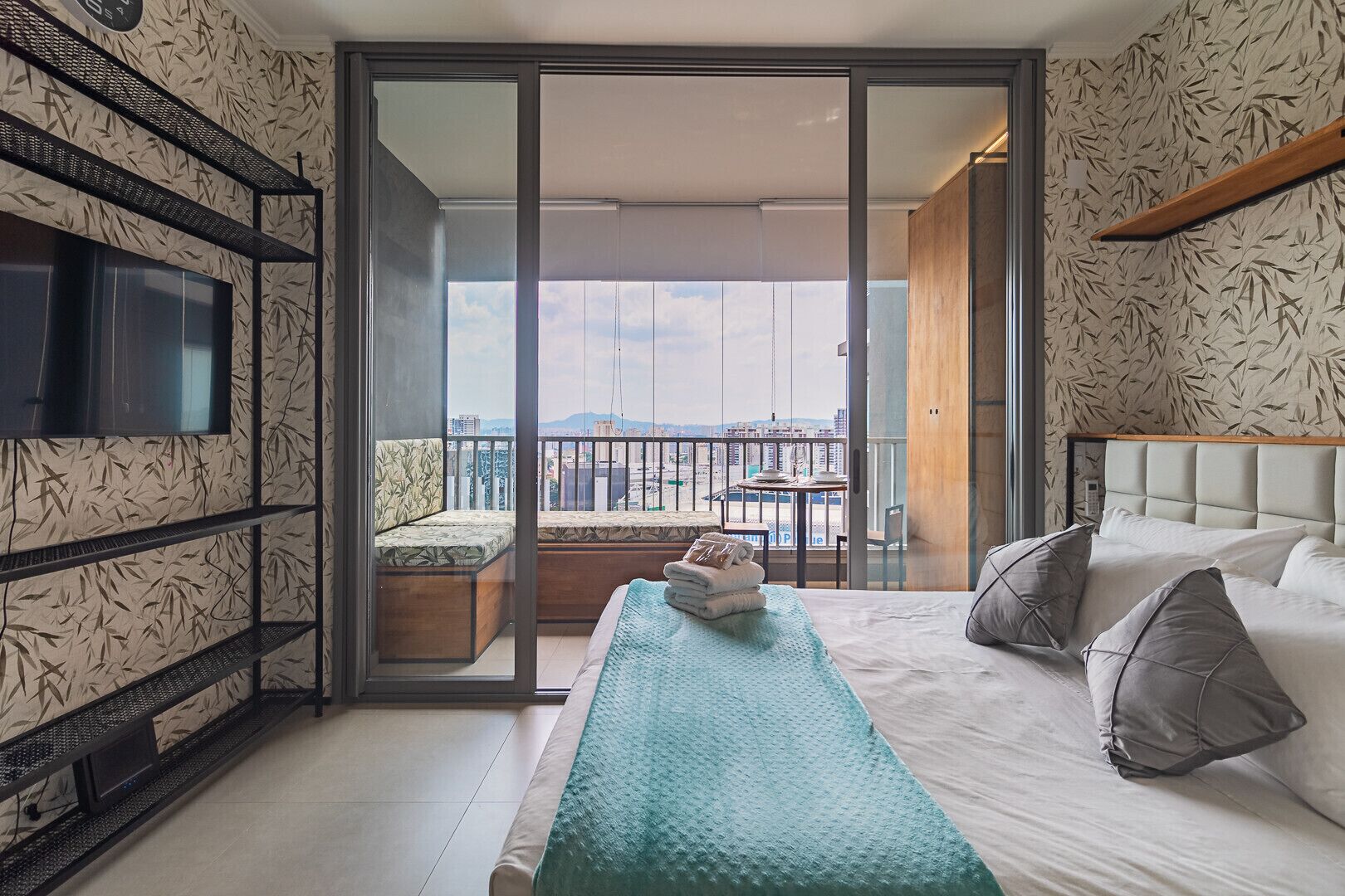 A modern hotel room with a balcony, a bed with pillows, a TV mounted on the wall, and a patterned wallpaper.