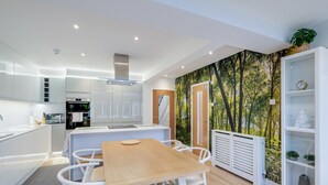 A modern kitchen with white cabinetry, a built-in oven, and a dining area with a wooden table and white chairs. - Bright 6 bedroom 3 bath house ideal for Pinewood Studios (Iver)