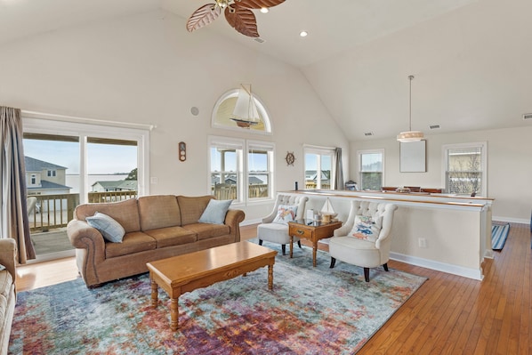 A spacious living room with a high ceiling, a large window overlooking a deck, a cozy seating area with a sofa and armchairs, and a wooden coffee table. - Sounds like a Plan: Pool | Hot Tub | Elevator | Dogs OK (Kill Devil Hills)