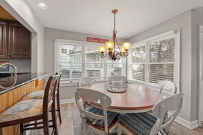A dining area with a wooden table, white chairs, a chandelier, and large windows with blinds. - Sea Rise: Beach Club Access w/ Pool | Golf Community (Nags Head)