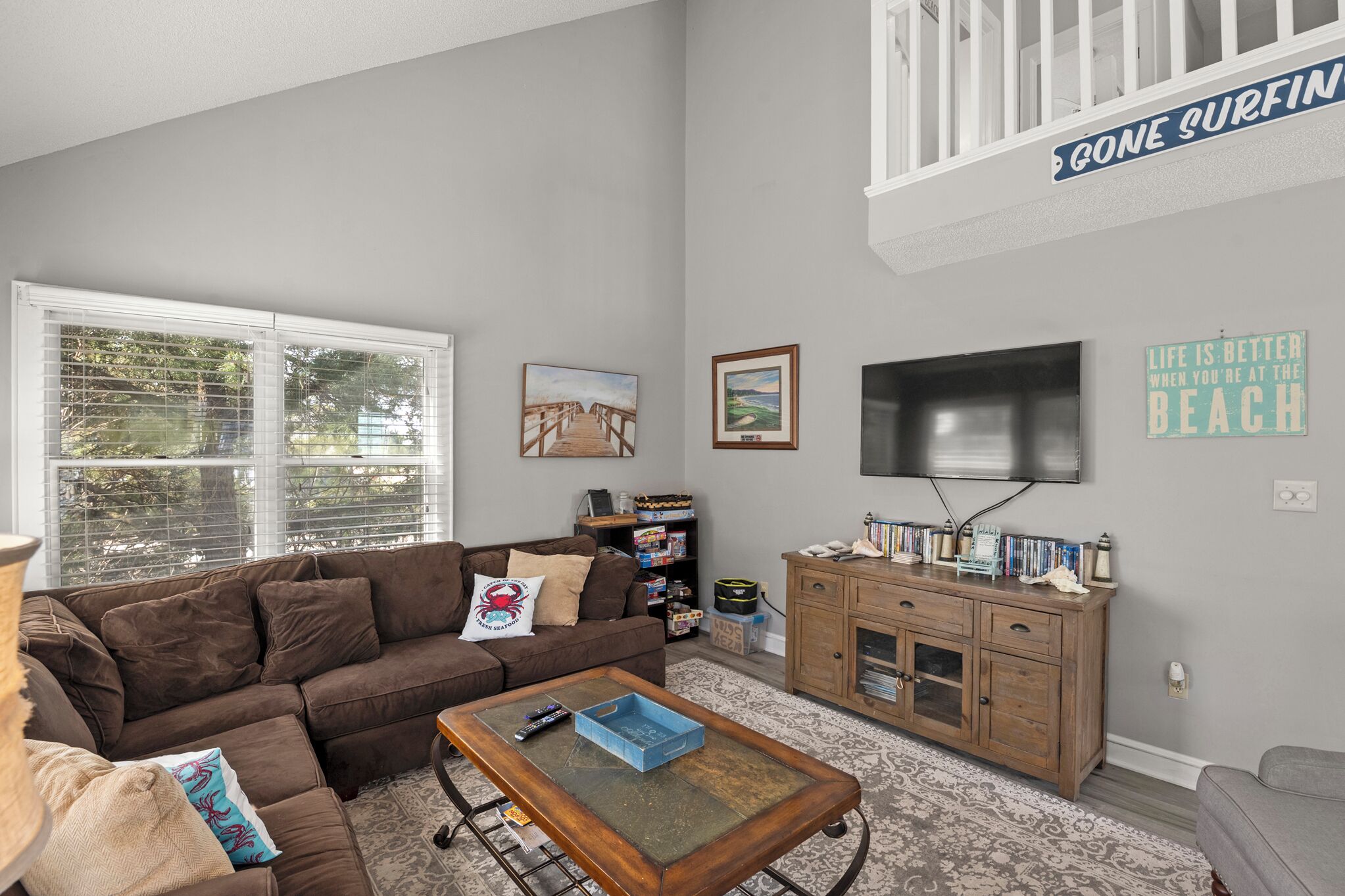 A living room with a brown sofa, a coffee table, a TV mounted on the wall, and a bookshelf.