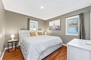 A bedroom with a large bed, a bedside table with a lamp, a window with a cityscape view, and a desk with a chair.