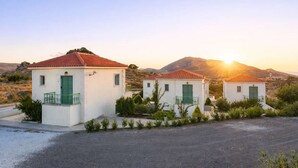 Three white buildings with red-tiled roofs and green shutters, surrounded by greenery and hills.
