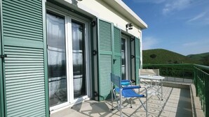 A balcony with a blue chair and a small table, a sliding glass door, and a view of green hills.