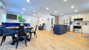 A modern kitchen with stainless steel appliances, a dining area with a blue sofa and black chairs, and a ceiling fan. - Sugarloaf Golf & Beach Retreat: Quiet Nature Edition (Cedar)