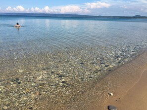 Une plage aux eaux claires et peu profondes, où l’on peut apercevoir des galets, et où une personne est en train de nager.
