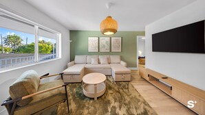 Un salon moderne avec un canapé d’angle, une table basse ronde, un téléviseur fixé au mur et une grande fenêtre donnant sur des palmiers.