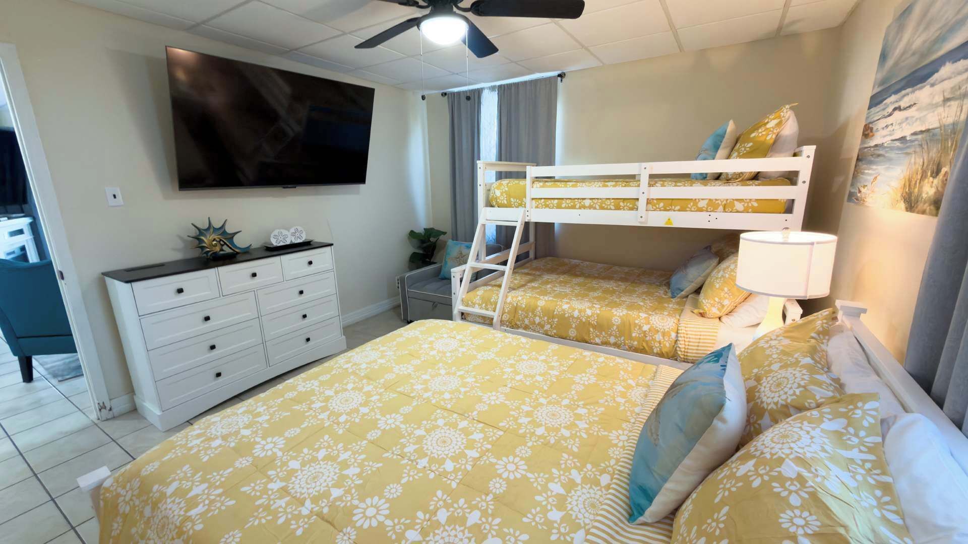 Une chambre avec un lit superposé, un téléviseur à écran plat, une commode et une fenêtre avec des rideaux.