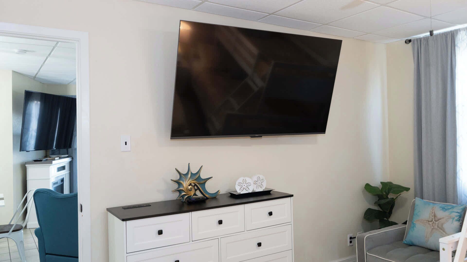 A living room with a flat-screen TV mounted on the wall, a white dresser with drawers, a blue chair, and a plant.