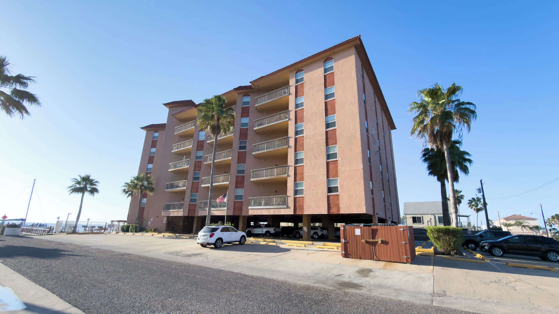 Un hotel de varios pisos con balcones, situado cerca de una calle con coches aparcados y palmeras.