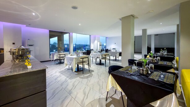 A spacious dining area with multiple tables, a kitchen area, and large windows offering a city view. - PEREGRINE HOTEL BY BOULEVARD (Jos)