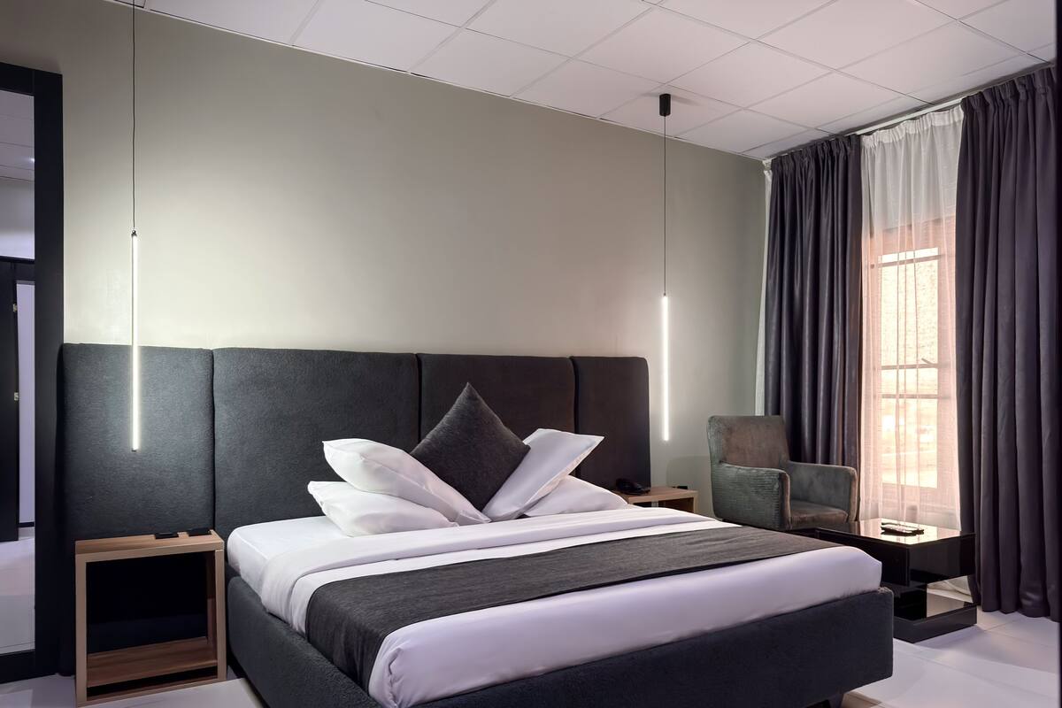 A modern hotel room with a large bed, bedside tables, a chair, and a window with curtains.