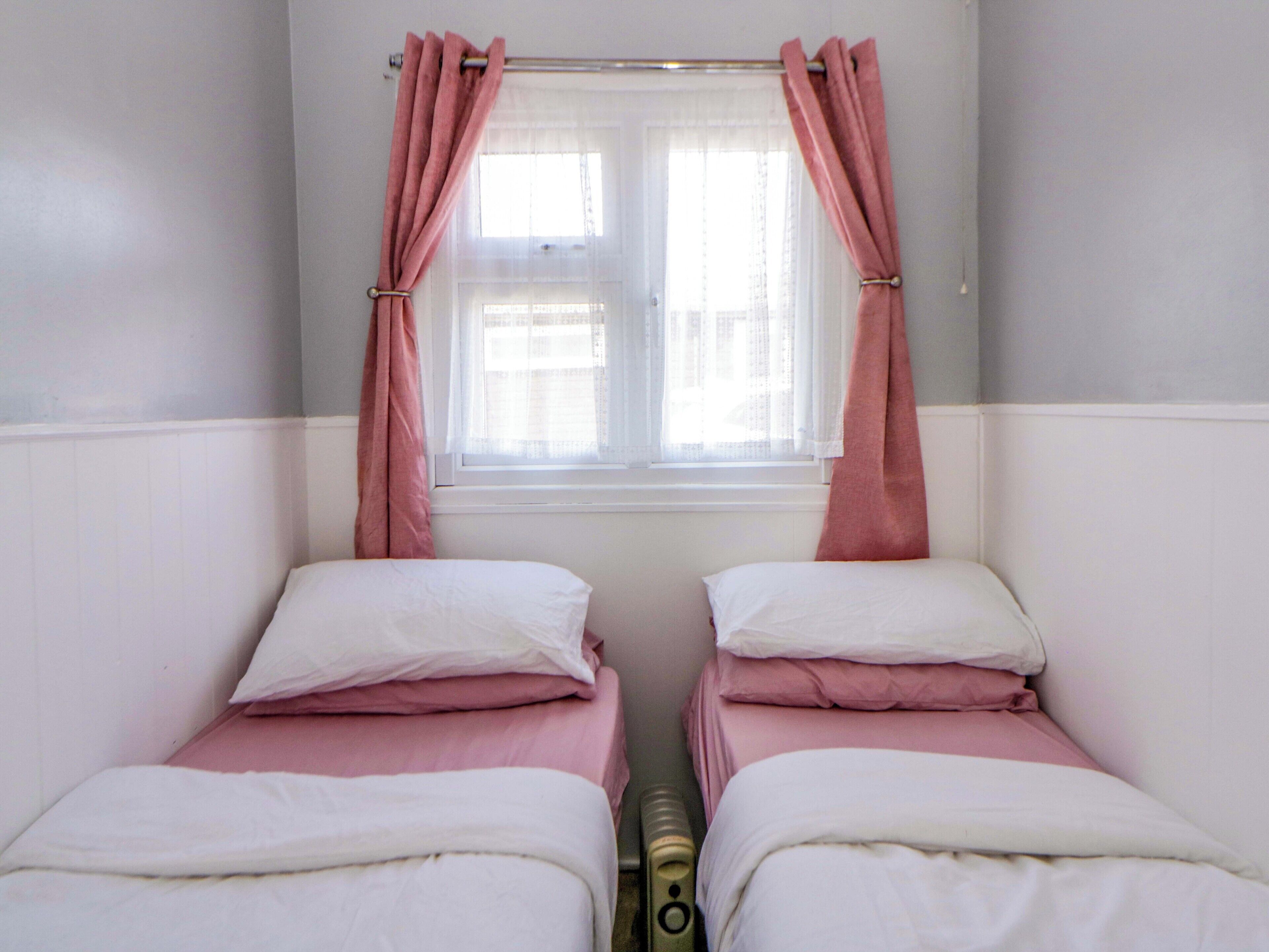 A room with two beds, each with white and pink bedding, a window with sheer curtains, and a radiator between the beds.