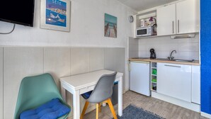 A compact kitchenette with a microwave, sink, and small refrigerator. There is a white table with a chair and a blue cushion.