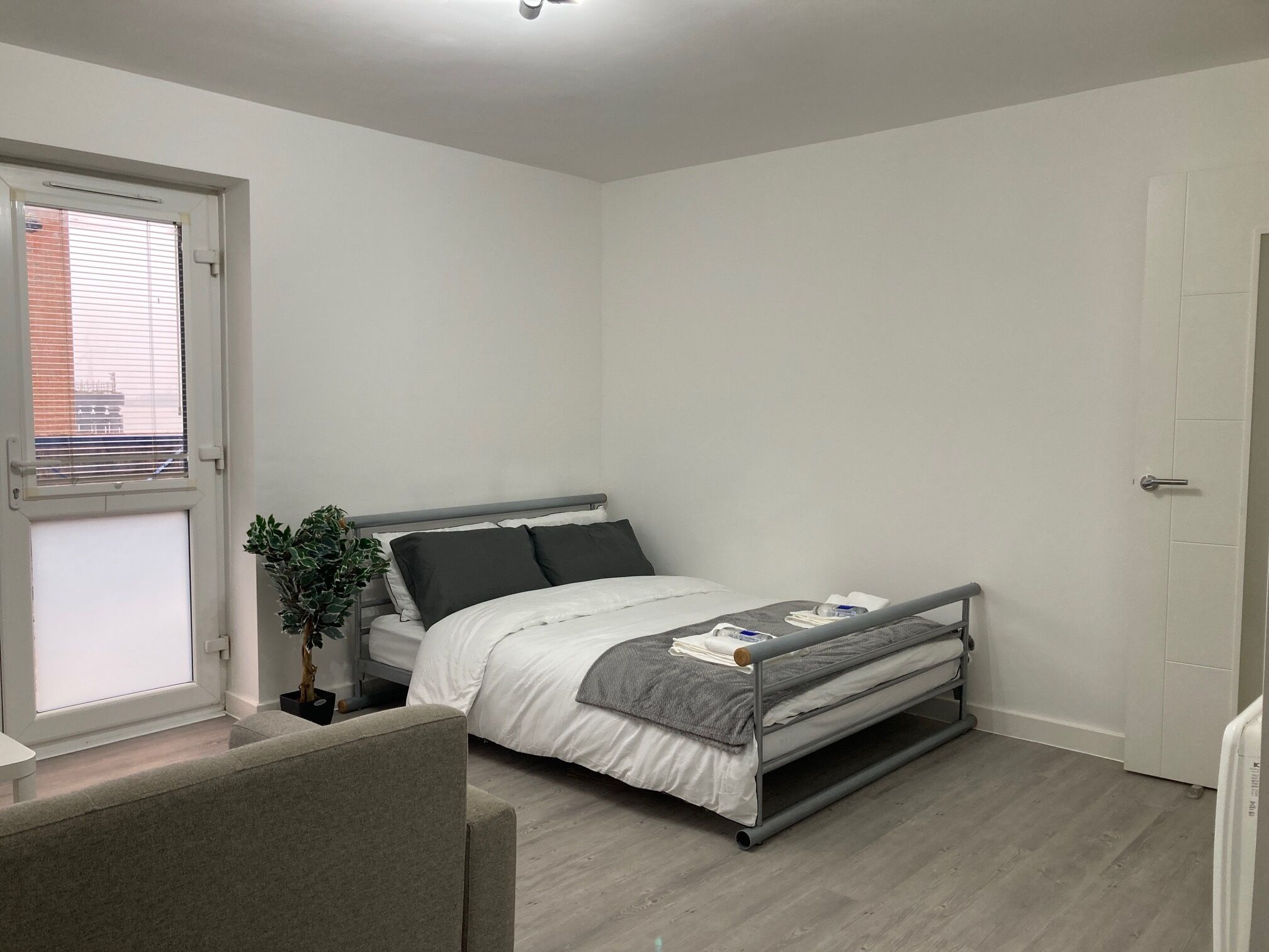 A compact bedroom with a bed, a window with blinds, a door, and a small plant.
