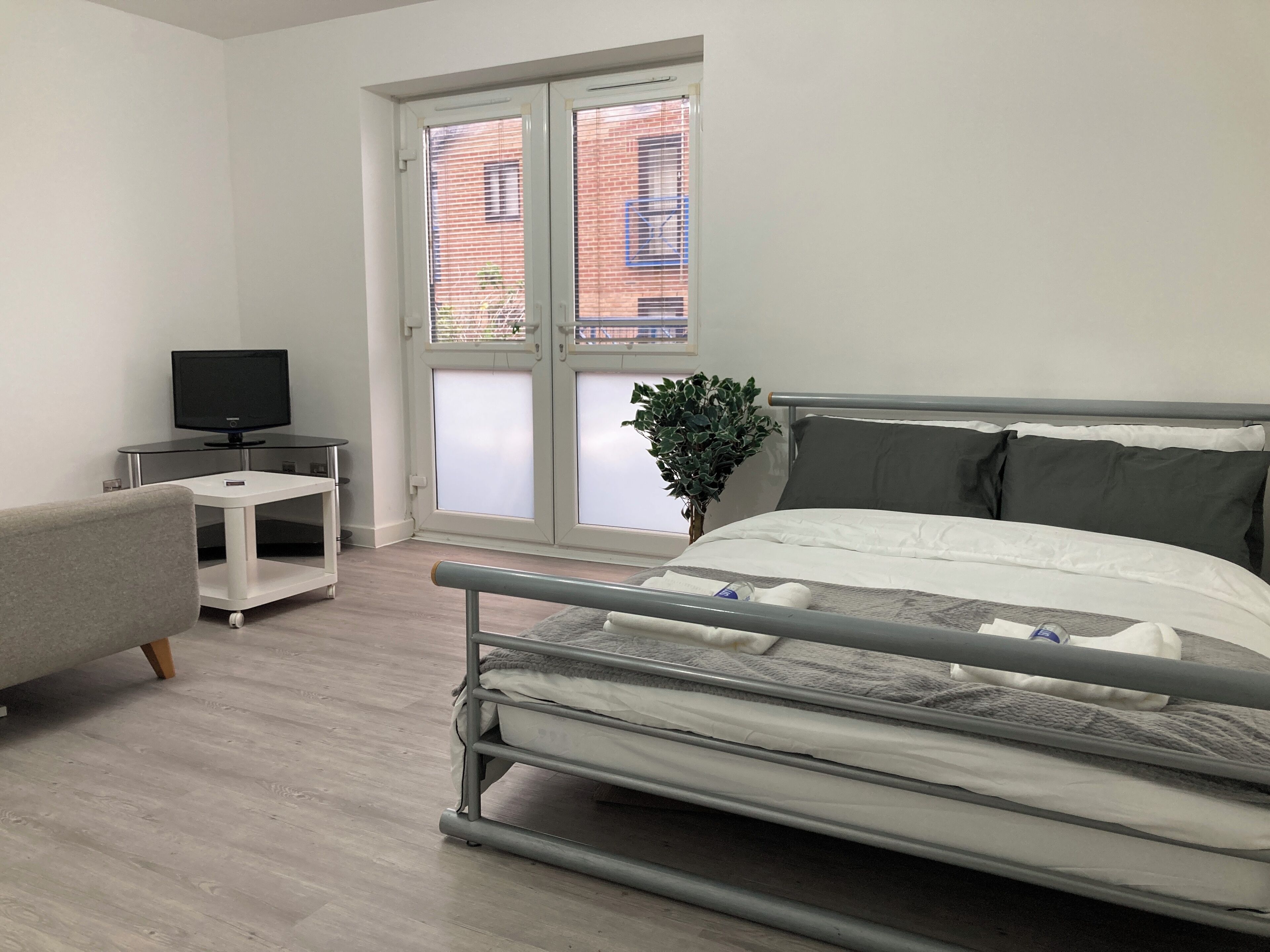 Ein modernes Schlafzimmer mit Bett, Fernseher auf einem Schrank, einer Topfpflanze und einem Fenster mit Jalousien.