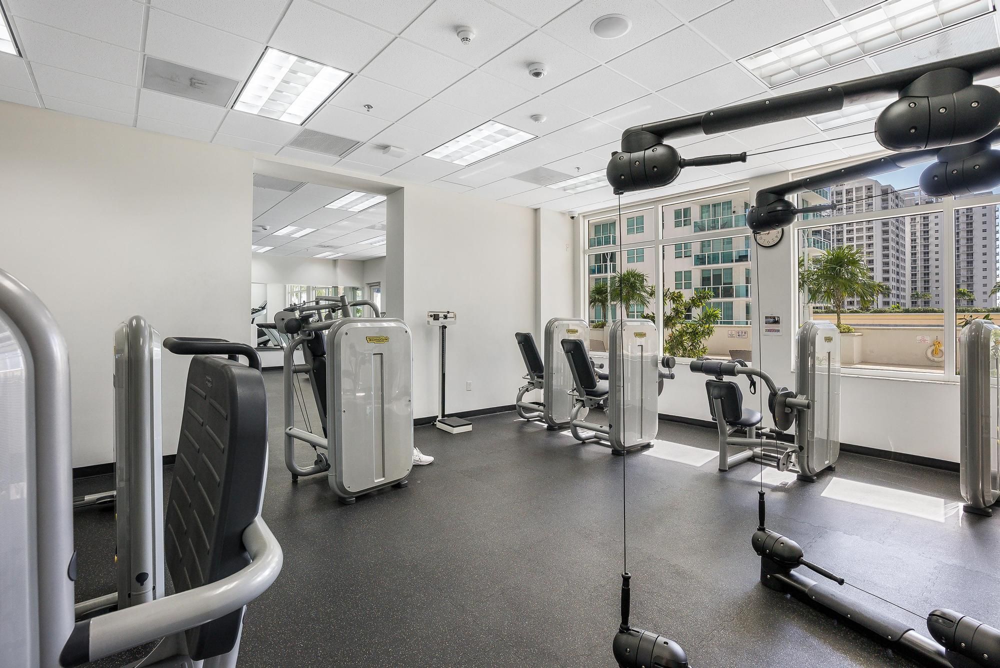 Un gimnasio con varias máquinas de ejercicio, ventanas amplias y vistas a edificios.