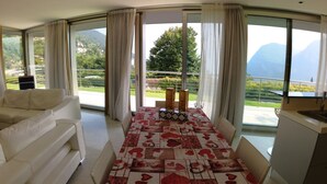 A room with a sofa, a table with a patterned tablecloth, a view of a balcony with a railing, and a mountain in the background. - A 5 minuti dal Lago di Tenno, spaziosa villa con piscina vista Lago di Garda (Trentino-Alto Adige)