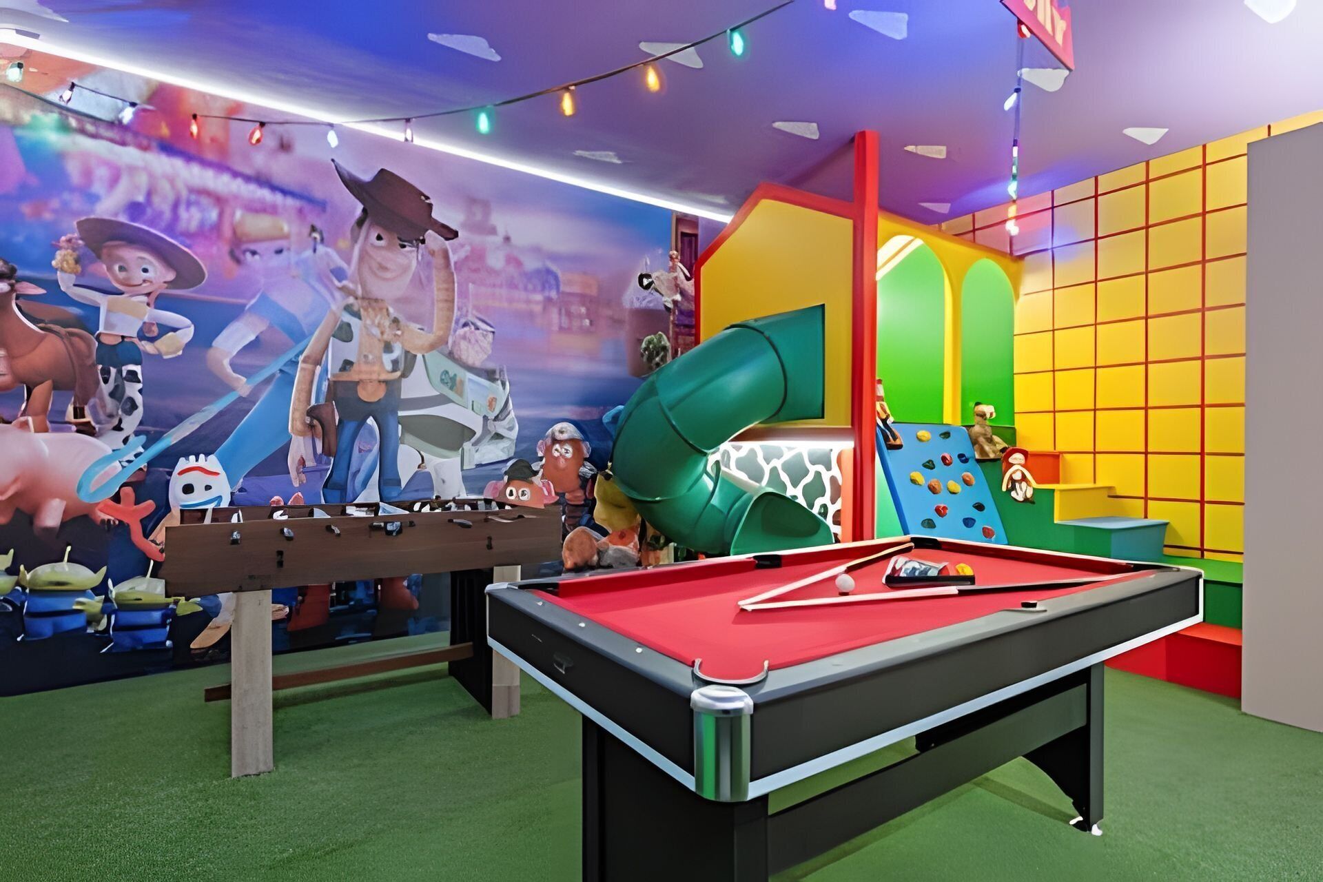 Zona de juegos infantiles con mesa de billar, estructuras de juego coloridas y un mural con personajes animados.