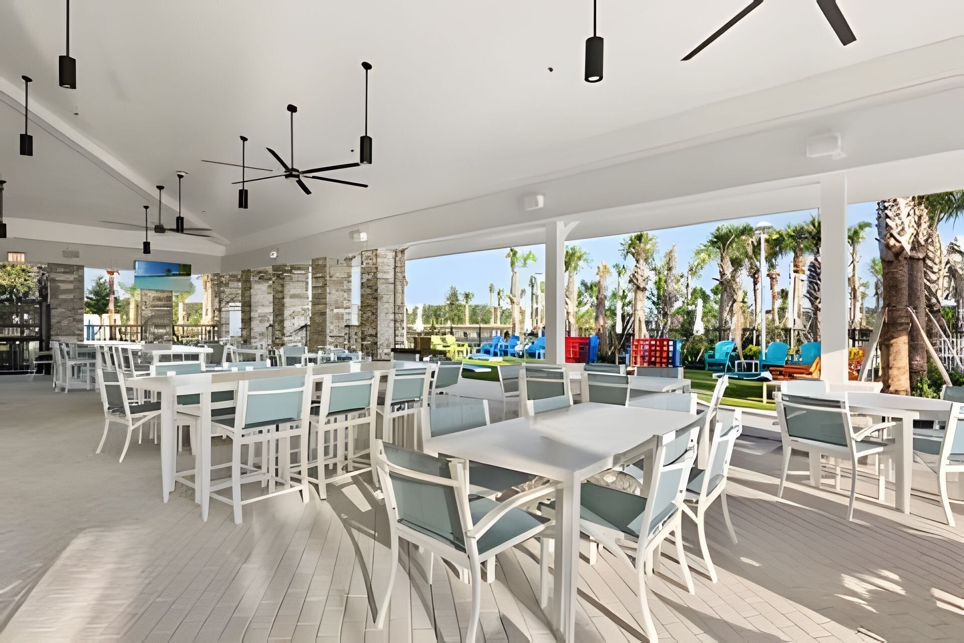 A spacious outdoor dining area with white tables and chairs, a ceiling fan, and a view of palm trees and a pool.