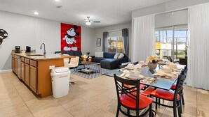 Un salon moderne avec une grande fenêtre, un coin repas avec des chaises rouges, une cuisine avec des meubles en bois et une œuvre d’art représentant Mickey Mouse accrochée au mur.