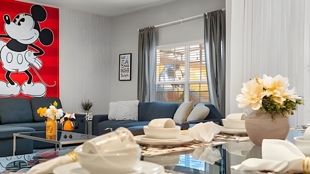 Un salon moderne avec un canapé bleu, une table basse sur laquelle se trouvent des assiettes et une théière, et une grande affiche de Mickey Mouse accrochée au mur.