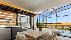 Une cuisine extérieure équipée d’un barbecue, d’une table et de chaises, avec vue sur une piscine et un jardin paysager.