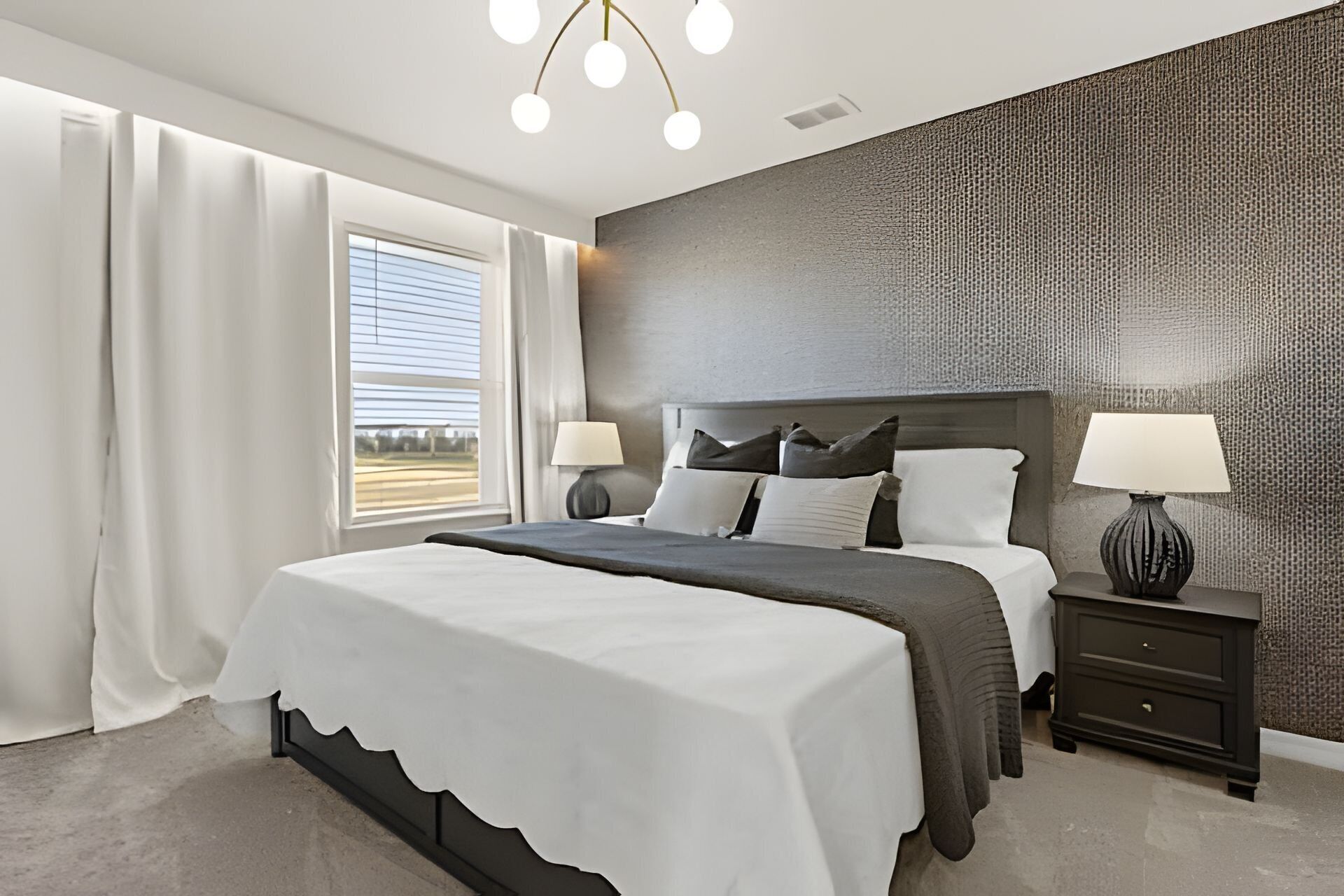 Ein modernes Schlafzimmer mit einem großen Bett, zwei Nachttischen, einer Wandlampe und Blick ins Freie durch ein mit Vorhängen drapiertes Fenster.