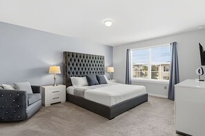 Une chambre à coucher moderne avec une tête de lit capitonnée, un lit recouvert de draps blancs, un canapé gris rembourré, deux tables de chevet avec des lampes et une grande fenêtre avec des rideaux.