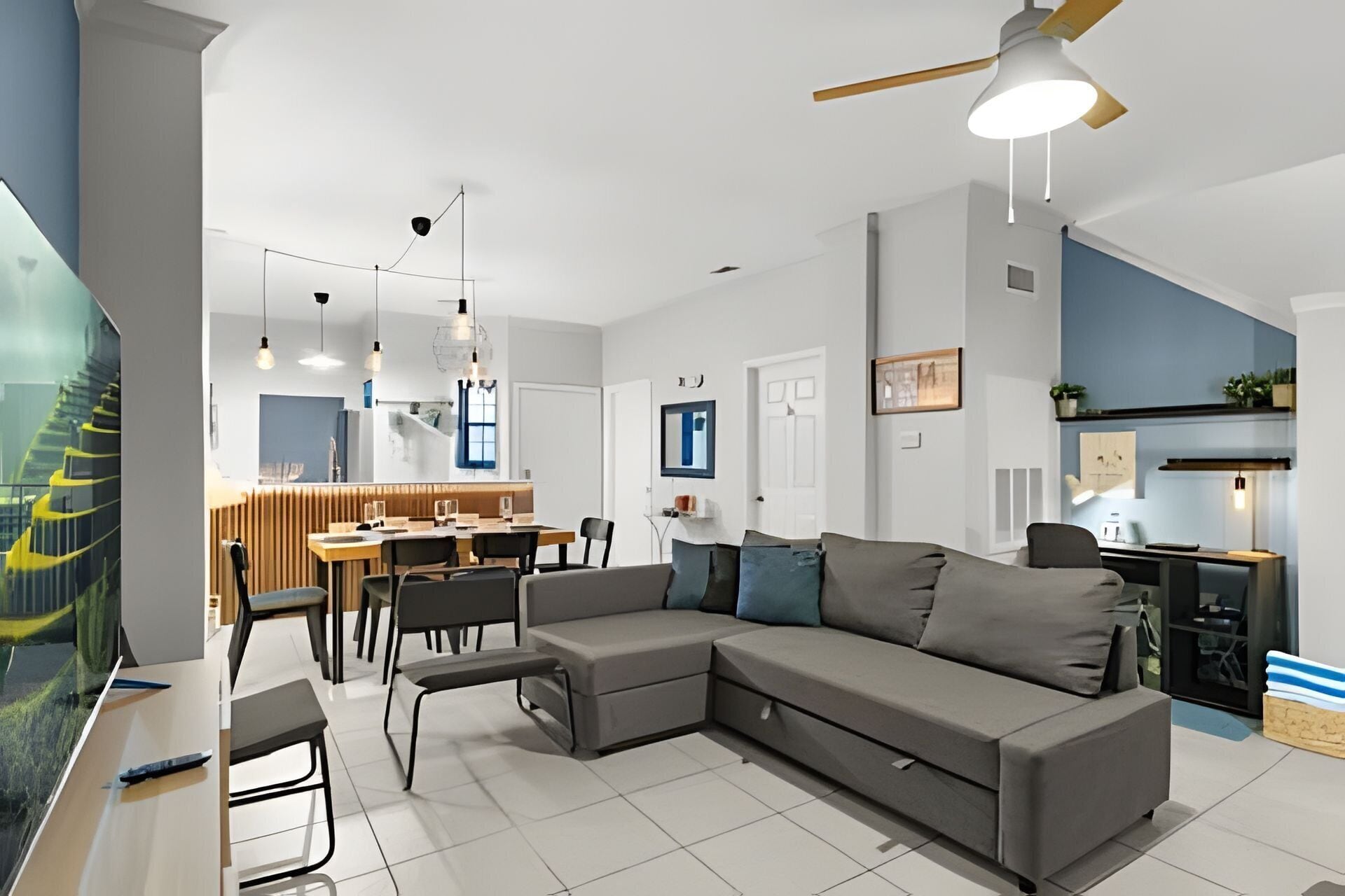 A modern living room with a grey sectional sofa, a dining area with a wooden table and chairs, a kitchen area with a countertop and appliances, and a ceiling fan.