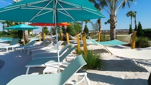 Een terras aan het strand met ligbedden en parasols, een zandstrand en palmbomen.