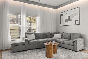 Un salon moderne avec un canapé d’angle gris, une table basse ronde en bois et une grande fenêtre agrémentée de rideaux blancs.