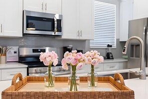 A kitchen with white cabinets, a microwave, and a stainless steel refrigerator. There are three glass vases with pink flowers on a wooden tray. - 5 Bd / 5 Ba / Sleeps 14 / Reunion Village (Kissimmee)