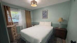 Ein Schlafzimmer mit Bett, Nachttisch, Lampe, Kommode, einem Fenster mit Vorhängen und einem gemusterten Teppich.