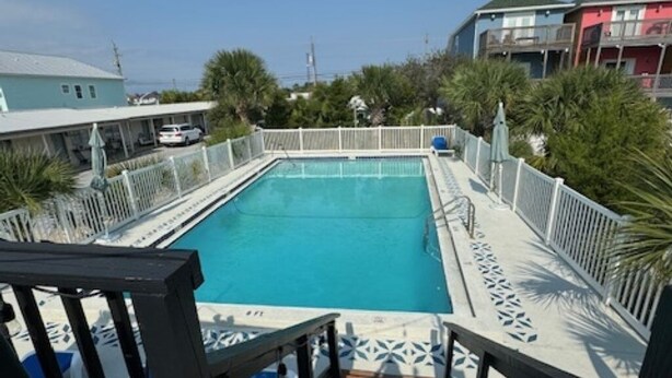 Ein rechteckiger Pool mit Holzterrasse und weißen Geländern, umgeben von Grünflächen und Gebäuden.