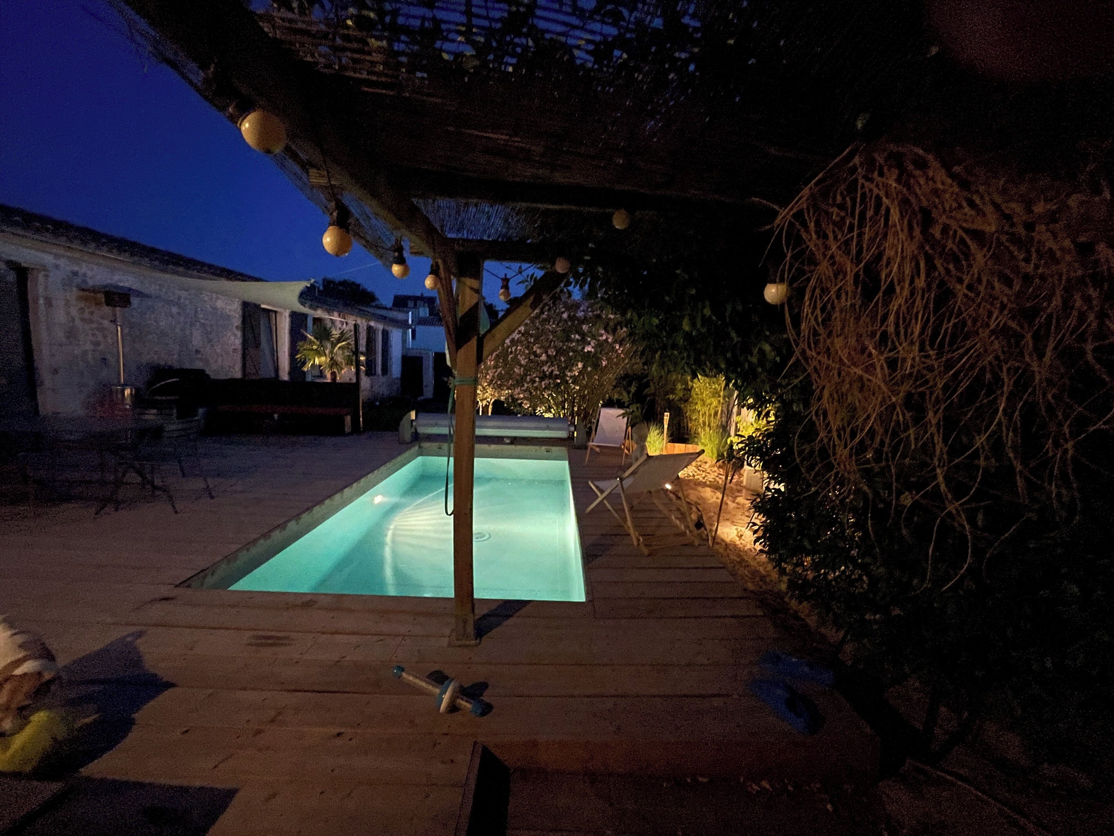 A rectangular pool with a wooden deck, surrounded by a pergola with hanging lights, a chair, and a small table.