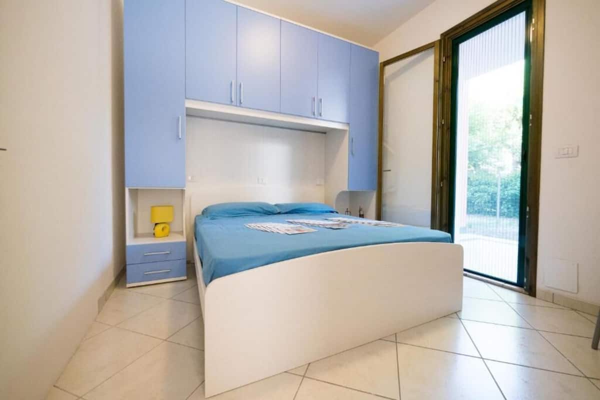 Un dormitorio con cama, mesita de noche, un armario azul y puerta corredera de vidrio al exterior.