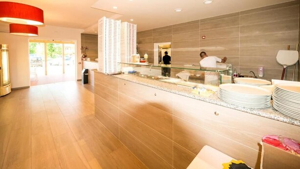 A modern hotel lobby with a glass partition, a reception area, and a dining area with stacked plates. - Pomposa Residence (Lido di Pomposa)