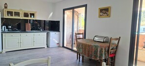 A compact kitchen with white cabinets, a black countertop, and a refrigerator. There's a dining table with a floral tablecloth and chairs. A painting is on the wall, and a window provides a view of the outside.