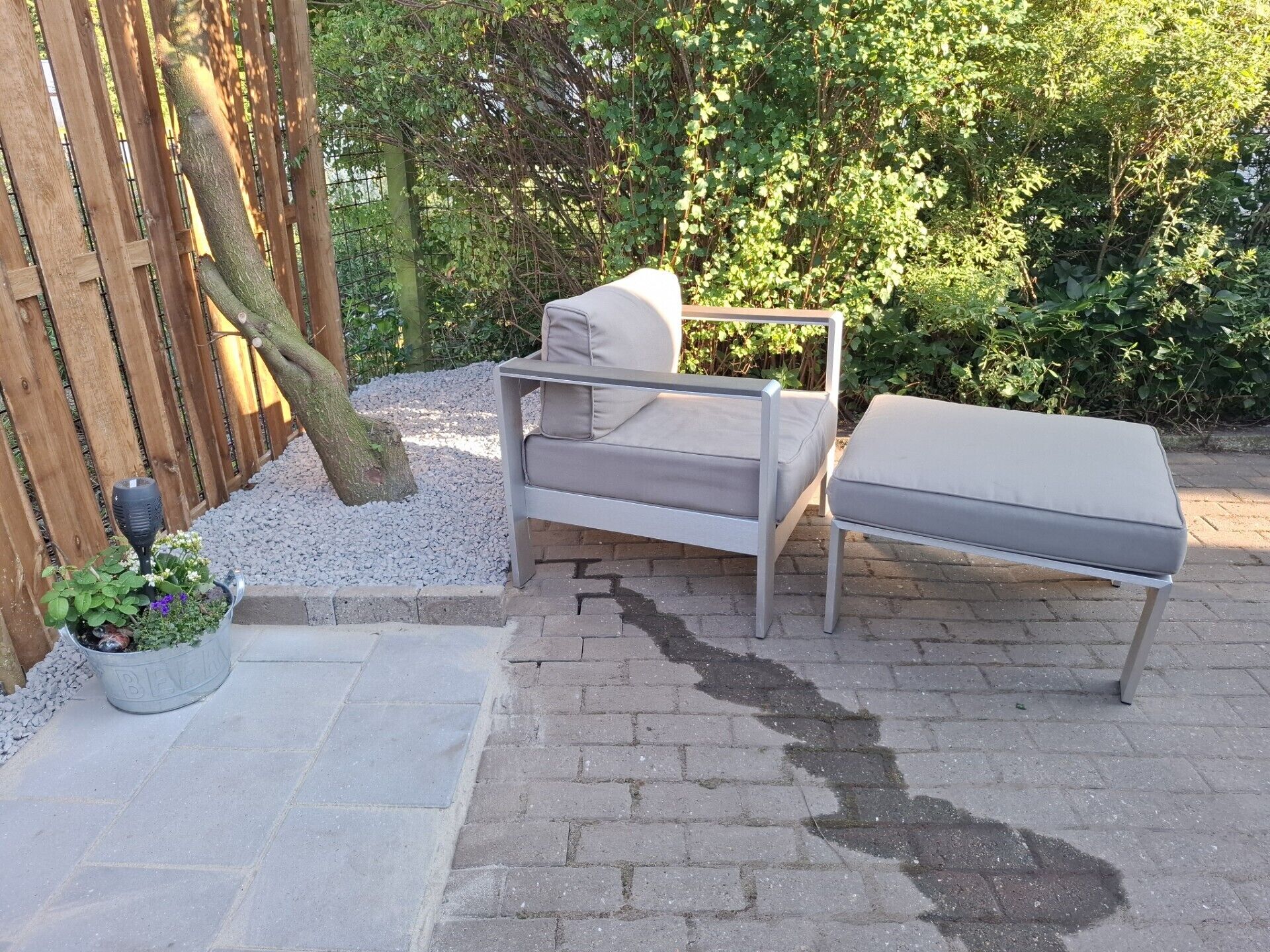 Eine Terrasse mit einer grauen Outdoor-Sofa und einer passenden Bank, umgeben von einem Holzzaun und viel Grün.