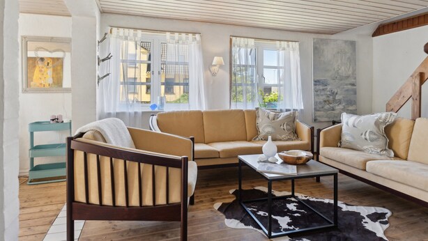 A living room with a sofa, a wooden chair, a coffee table, and a cowhide rug. There are windows with curtains and a painting on the wall. - The Cozy Cottage – Charming Holiday Home near the North Sea (Hirtshals)