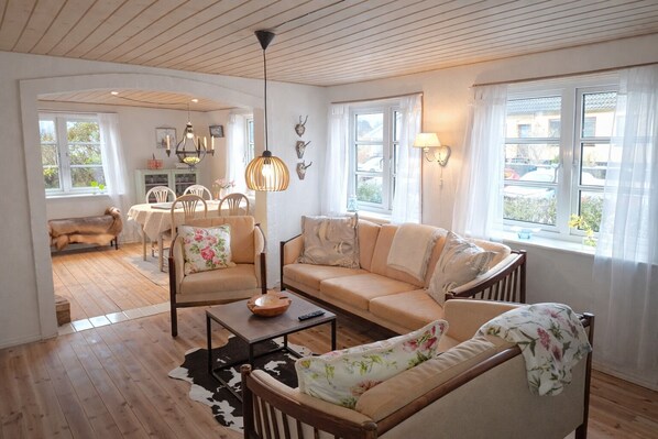Un salon chaleureux avec un canapé, des fauteuils, une table basse et, au fond, un coin repas.