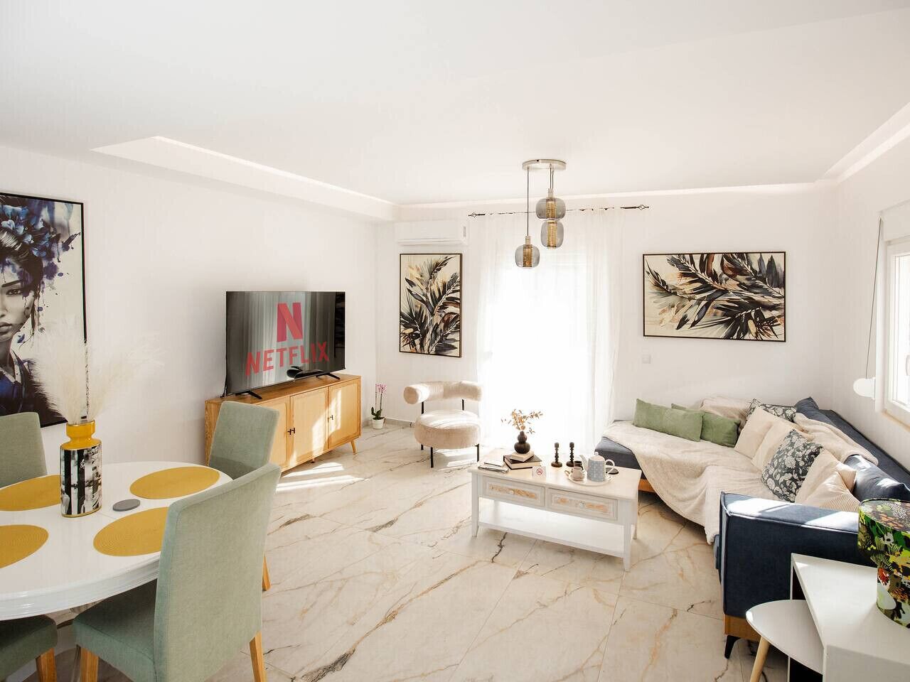 Ein modernes Wohnzimmer mit einem weißen Sofa, einem Esstisch mit gelben Untersetzern, einem Fernseher auf einer Holzkommode und Kunstwerken an den Wänden.