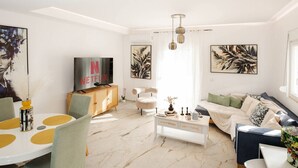 A modern living room with a white sofa, a dining table with yellow placemats, a TV on a wooden cabinet, and artwork on the walls. - Spacious Gournes Home for 7 | 2.5 Baths (Gournes)