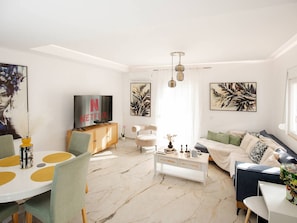 Ein modernes Wohnzimmer mit einem weißen Sofa, einem Esstisch mit gelben Untersetzern, einem Fernseher auf einer Holzkommode und Kunstwerken an den Wänden.