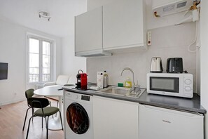 A compact kitchen with a washing machine, a microwave, a kettle, and a toaster. There is a dining table with chairs and a window with blinds. - Bail mobilité - Passy - Studio - Trocadéro (Paris)