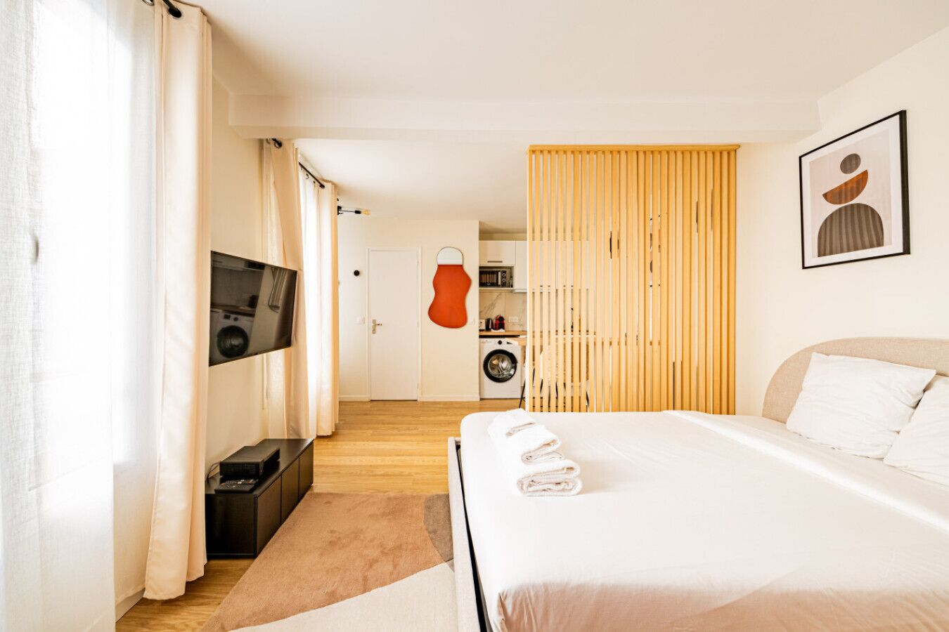 A modern hotel room with a large bed, a flat-screen TV mounted on the wall, a washing machine, and a wooden partition.