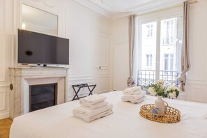 A hotel room with a large bed, a flat-screen TV, a fireplace, a balcony view, and a wicker basket with towels. - Appartement 4 Chambres/4 SDB/Paris 17 (Paris)