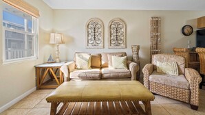 A living room with a beige sofa, a wicker armchair, a wooden coffee table, and two decorative wall pieces. - ✨Beachfront Complex + Community Pool | Sleeps 4 | Sunrise Village 111✨ (Gulf Shores)